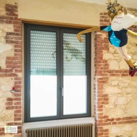 Serramento a due ante in alluminio scuro su muro di pietra chiara con pupazzo in cartapesta che suona il violino in appartamento ad Augusta